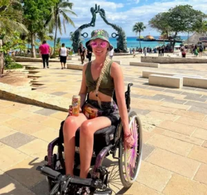 A woman in a wheelchair enjoys a sunny day at a tropical plaza near the coast. She is wearing a green crop top, a floral hat, and pink heart-shaped sunglasses, holding a drink. Behind her is a large bronze sculpture of two figures reaching toward each other, with turquoise water and palm trees in the distance.