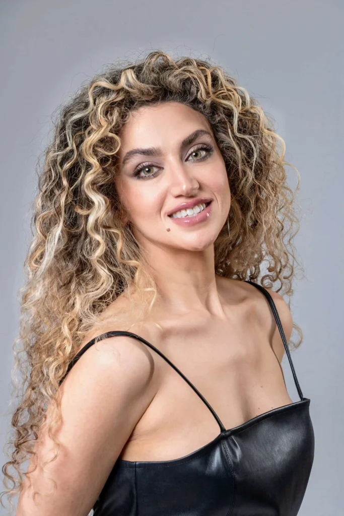 A close-up headshot of a woman with large, light-colored eyes and voluminous curly hair. she is smiling at the camera and wearing a black spaghetti-strap top.