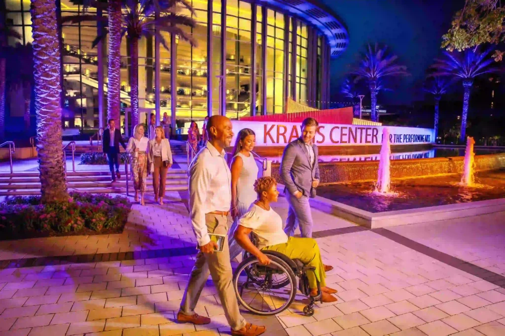 Group of visitors, including a wheelchair user, walking along a well-lit outdoor plaza at the Kravis Center for the Performing Arts at night, with palm trees, fountains and the glass-fronted theatre building illuminated in purple and gold, highlighting an accessible arts and cultural destination.