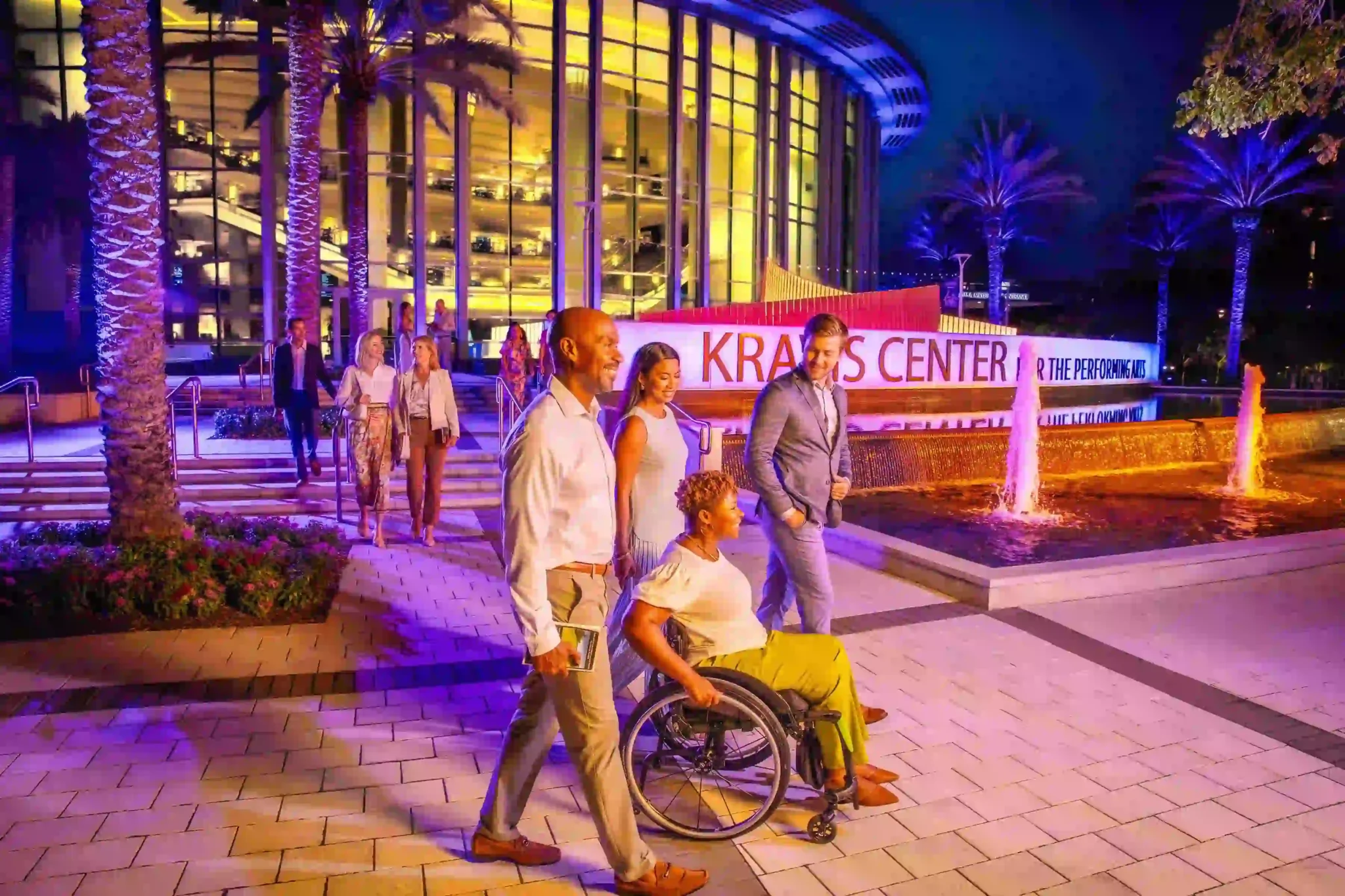 Group of visitors, including a wheelchair user, walking along a well-lit outdoor plaza at the Kravis Center for the Performing Arts at night, with palm trees, fountains and the glass-fronted theatre building illuminated in purple and gold, highlighting an accessible arts and cultural destination.