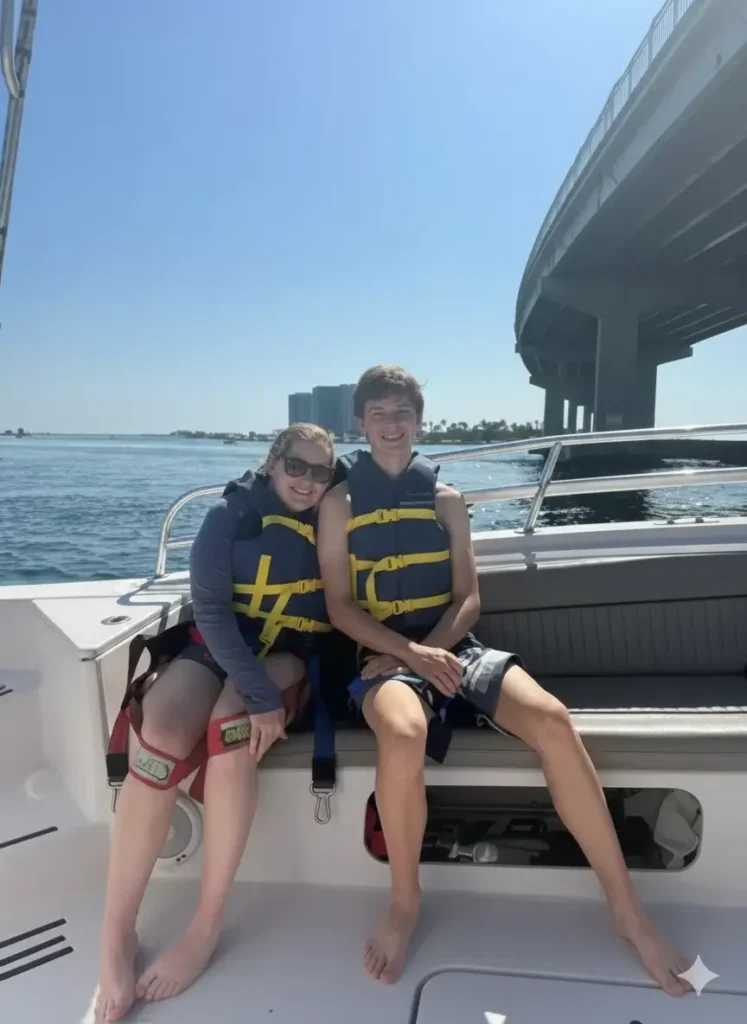 A teenage boy and a woman sitting side-by-side on the deck of a boat under a bright blue sky. Both are wearing yellow and blue life jackets and smiling. A large concrete bridge spans the water in the background.