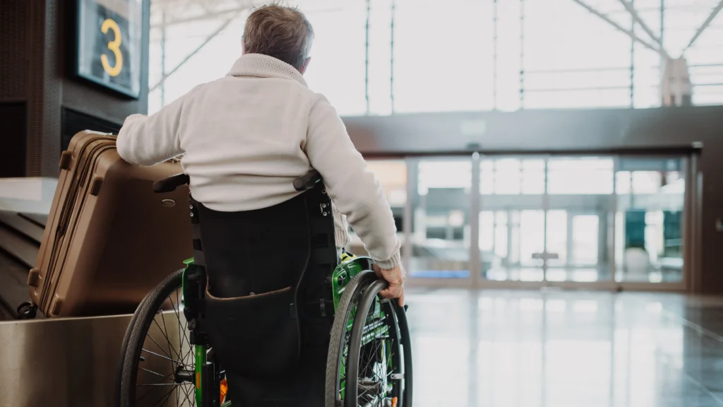 Rear view of a man in a white sweater sitting in a green wheelchair at an airport terminal, looking toward the departure gates with a large brown suitcase beside him.