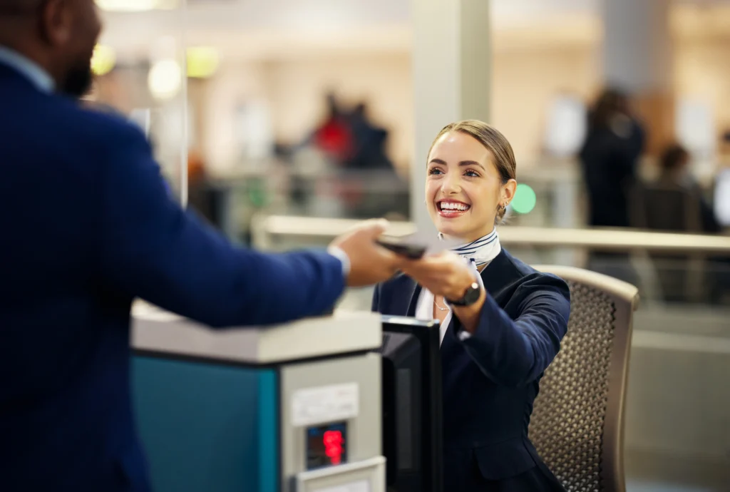 A smiling female airport service agent in a navy uniform and neck scarf handing a passport and boarding pass back to a passenger at a check-in counter.