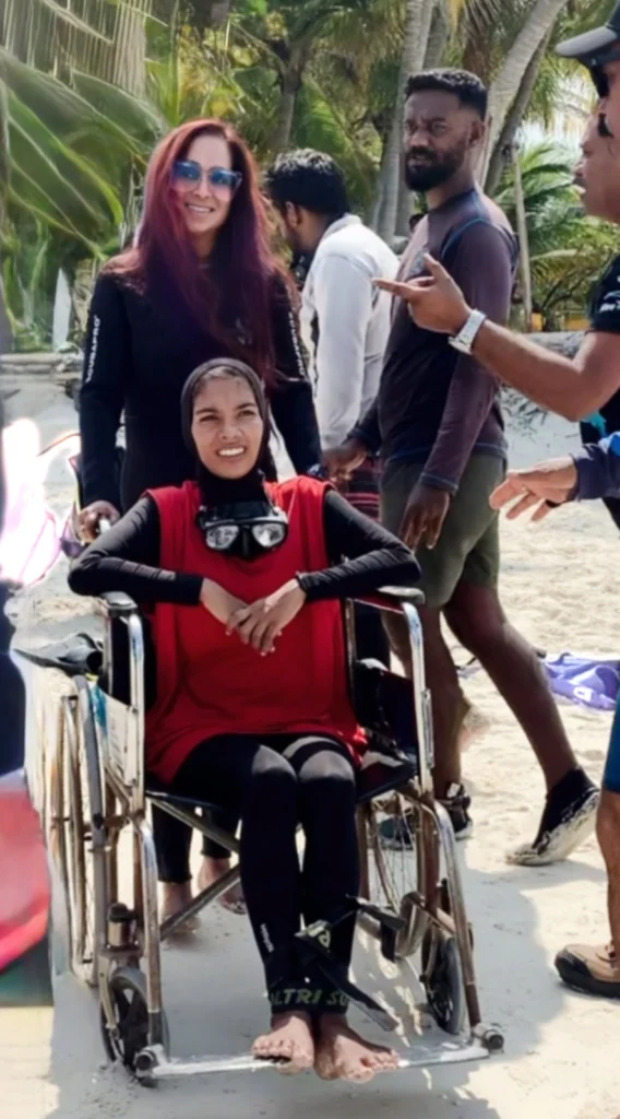 A woman seated in a wheelchair on a sandy beach, wearing a red vest over a black long-sleeved swim shirt and a diving mask around her neck. Behind her, a woman with long reddish-brown hair and sunglasses smiles at the camera, while other people and palm trees are visible in the background.