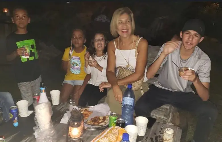 A candid nighttime photo of a woman smiling with four children gathered around a rustic wooden table. The table is cluttered with snacks, drinks, and a glowing lantern, creating a warm, social atmosphere.
