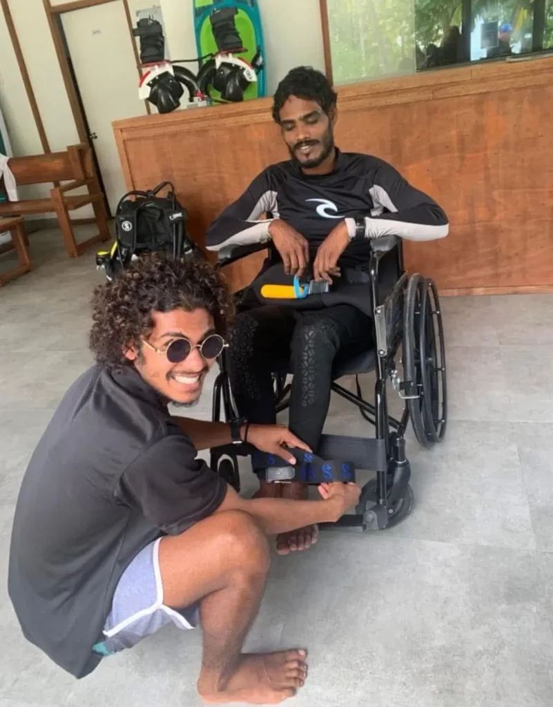 A man with curly hair and sunglasses kneeling to adjust a strap on the leg of another man seated in a wheelchair. Both are in a dive shop or seaside facility, with scuba gear and a wooden counter in the background.