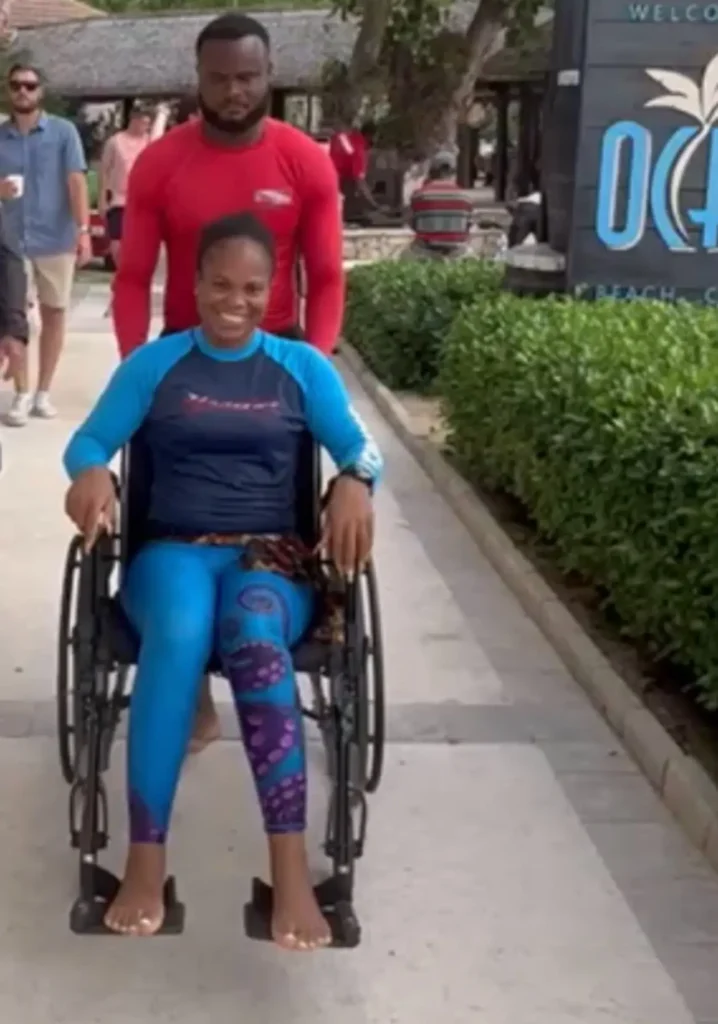 A woman in a blue long-sleeved swim shirt and patterned leggings smiles while sitting in a wheelchair. A man in a red long-sleeved shirt is standing behind her, pushing the wheelchair along a paved path at an outdoor tropical resort.