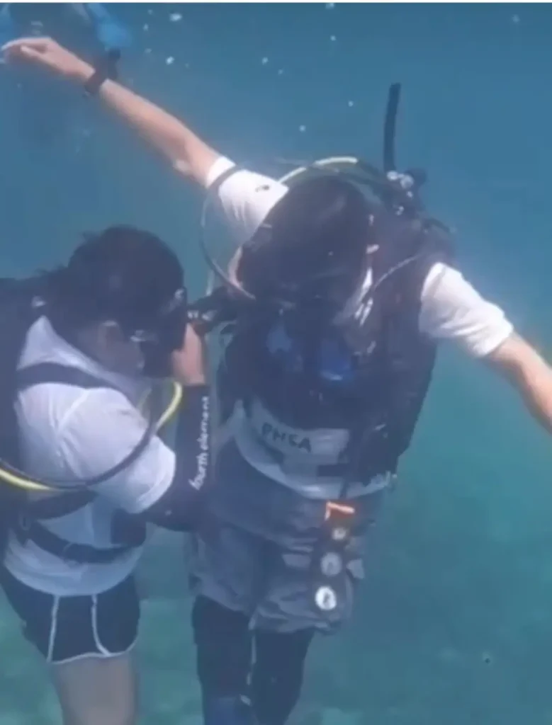 An underwater shot of two scuba divers. One diver is assisting the other with their gear, specifically checking the chest area of the buoyancy control device (BCD) while submerged in clear blue water.