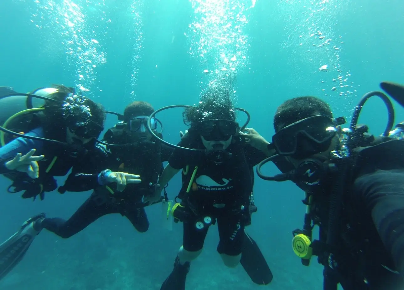 An underwater group selfie of four scuba divers. They are wearing full gear, including masks and regulators, and are posing with hand signals while bubbles rise around them in the bright, turquoise ocean.