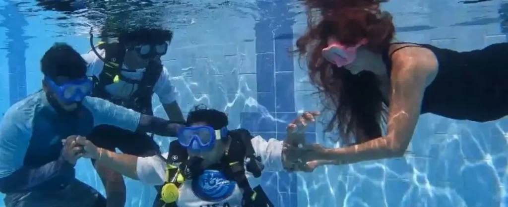 A group of four people practicing scuba skills in a swimming pool. Three individuals are wearing scuba tanks and masks, while a fourth person in a black swimsuit and pink mask swims alongside them, assisting a diver in the center.