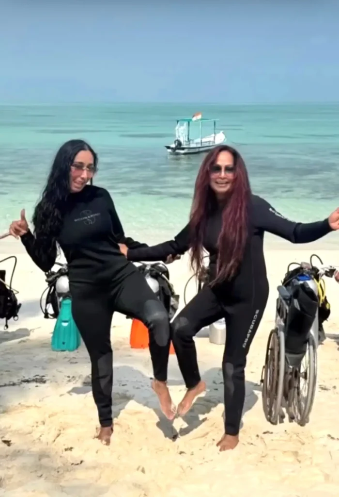 Two women in black wetsuits posing playfully on a sunny white-sand beach. They are standing on one leg with their feet touching. Scuba tanks and a wheelchair are visible on the sand behind them, with a small boat floating in the calm, tropical water in the background.