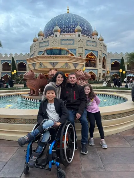A family of five poses for a portrait in front of a grand, ornate building with a large blue dome and intricate patterns. In the foreground, a young boy sits smiling in a blue wheelchair. Behind him, two adults and two other children are grouped together around the edge of a large, circular stone fountain with animal-shaped water spouts.