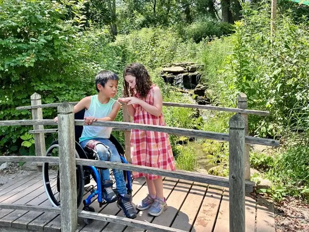 A young boy in a wheelchair and a young girl in a red plaid dress stand on a wooden bridge in a lush, green park. They are both leaning in to look closely at something the girl is holding in her hands. In the background, a small, tiered rock waterfall flows into a stream surrounded by dense foliage.
