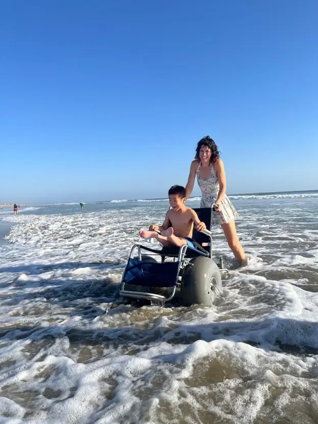 A woman in a floral dress pushes a young boy in a specialized beach wheelchair through the shallow, foamy surf at a beach. The boy is laughing with his arms up as the water splashes around the large, grey balloon tires of the chair. The sky is a clear, bright blue above the ocean horizon.