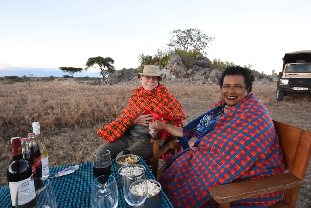 A smiling couple sits at a table in a grassy field during a safari sundowner. They are wrapped in vibrant red and blue plaid Maasai blankets. The table is set with snacks, wine, and glass-bottled sodas, with a safari vehicle parked in the background under a pale evening sky.