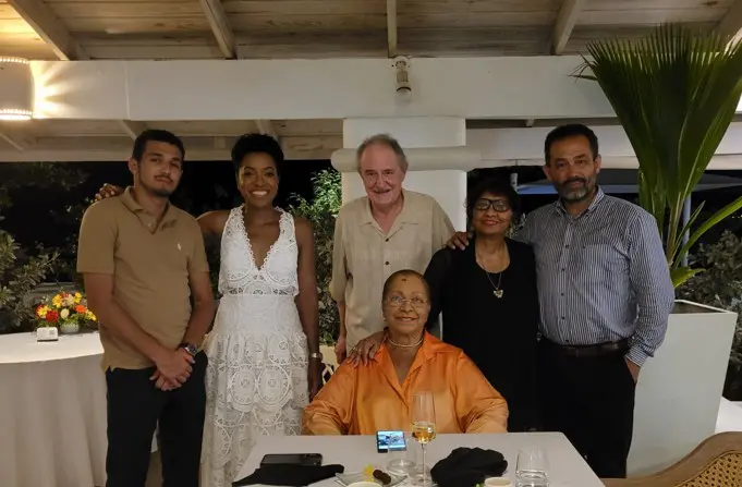 A formal group photo of six people posing together at a restaurant. A woman in an orange silk blouse is seated at the center of the table, surrounded by five standing adults under the soft lighting of a covered patio.