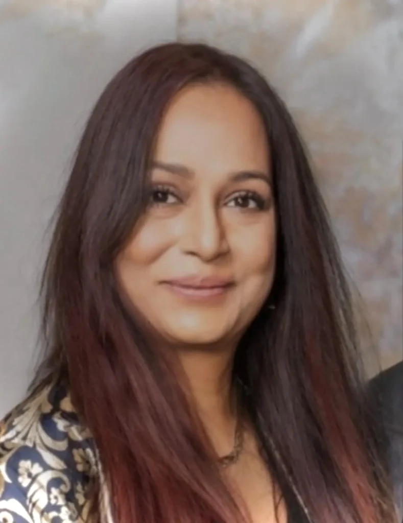 A close-up portrait of a woman with long, dark hair featuring reddish-brown highlights. She is smiling gently at the camera, wearing a gold necklace and a patterned garment with gold and navy blue designs.