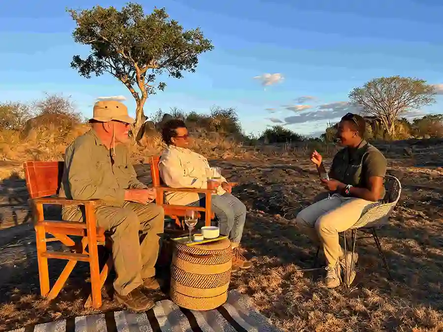Three people sitting outdoors in wooden chairs at golden hour, engaged in conversation around a small woven basket table with drinks. They're on a wooden deck in a savanna landscape with scattered trees and bushes under a blue sky with light clouds. The person on the left wears a khaki hat and jacket, the person in the middle wears a light-colored outfit, and the person on the right wears a dark green shirt and khaki pants, gesturing while talking