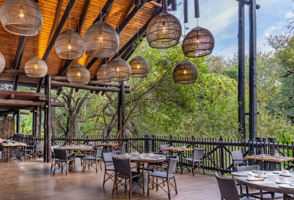An open-air restaurant deck with wooden flooring and a high, thatched-style roof. Several round wooden dining tables are set with white linens, surrounded by grey wicker chairs. Large, spherical woven pendant lights hang from the rafters, and the deck overlooks a lush green forest.