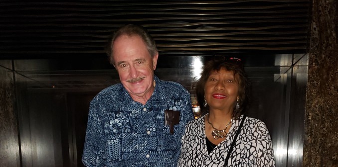 A man and woman posing together in front of a modern fireplace. The man is wearing a blue patterned shirt and khaki trousers, while the woman is in a black and white graphic print top.