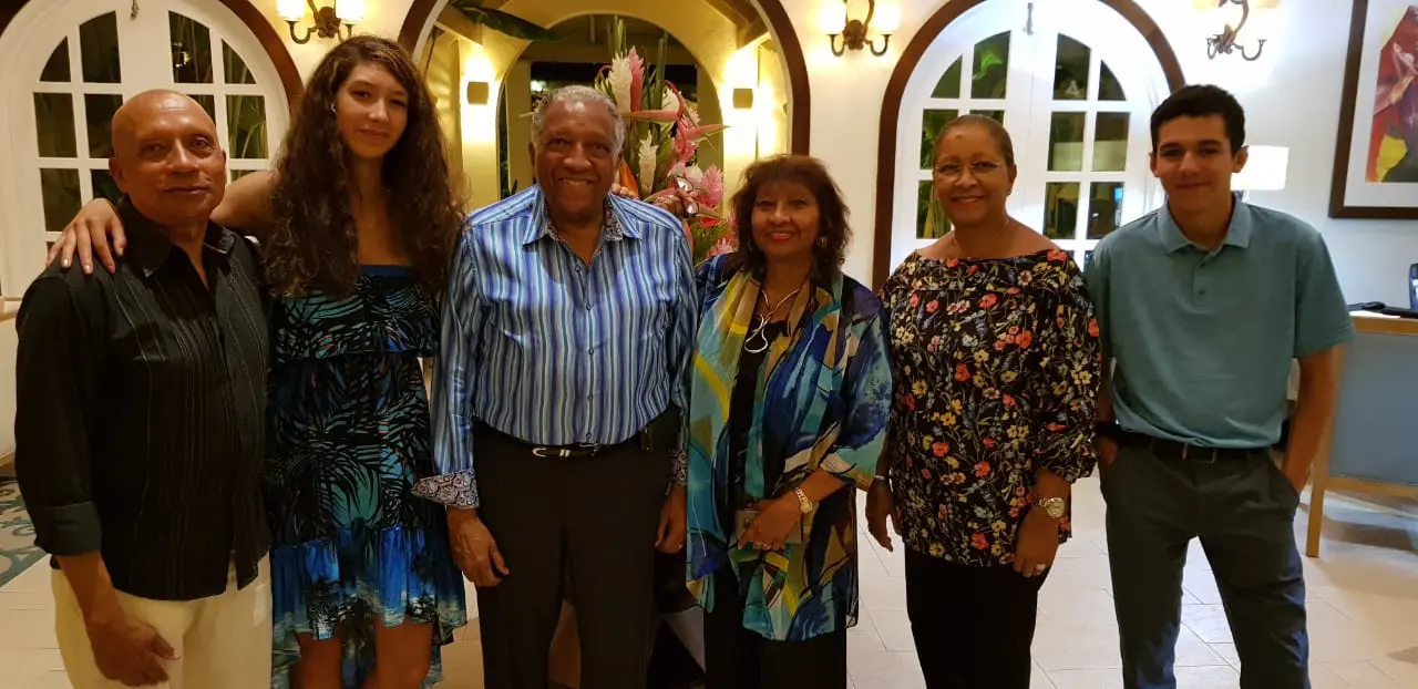 Six people posing for a photo in an elegant indoor hallway with arched doorways. They are dressed in a mix of colorful patterns and smart-casual attire, standing in a line in front of a large floral arrangement.