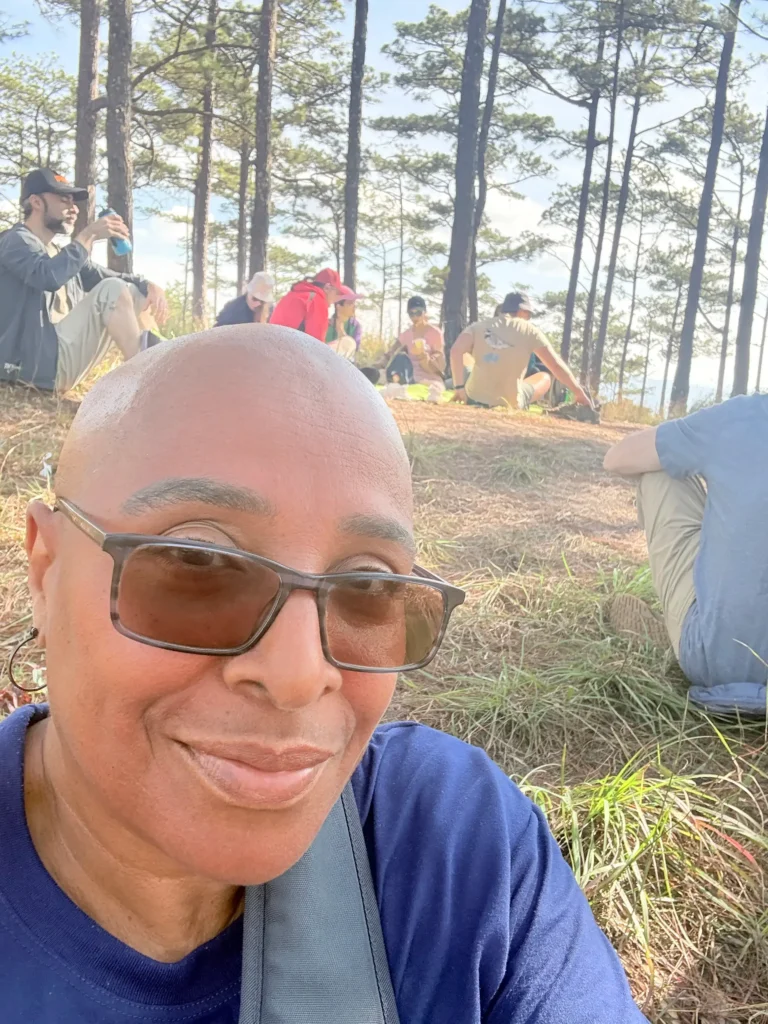 A close-up selfie of a person with a shaved head, smiling warmly while, wearing tinted glasses and a blue shirt. They are outdoors in a sunlit pine forest. In the background, a small group of people is sitting on the grassy ground, resting among the tall trees.