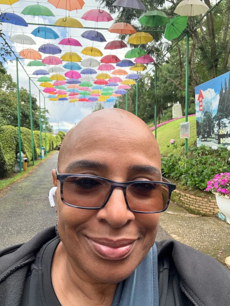 A close-up selfie of a smiling person with a shaved head and tinted glasses. They are standing on a paved path beneath a long canopy decorated with dozens of colorful, hanging umbrellas in shades of yellow, pink, blue, and green. Lush green bushes and trees line the path in the background.