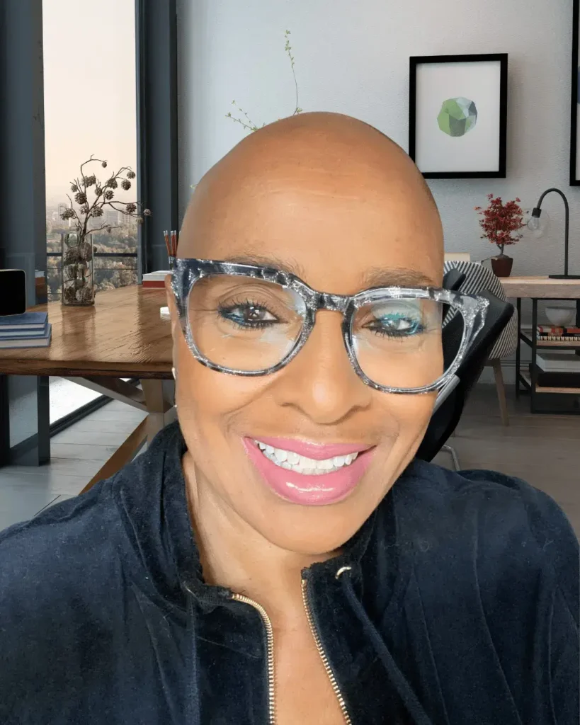 A close-up selfie of a person with a shaved head, smiling warmly while wearing stylish, thick-rimmed clear glasses. The background is a modern, bright office or study with a wooden desk, a minimalist lamp, and a large window overlooking a city view at dusk.