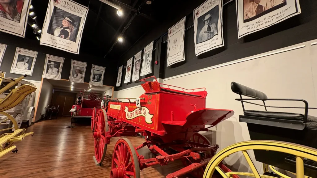 An indoor museum gallery featuring vintage horse-drawn vehicles. In the foreground is a bright red "Alert Hose Co." fire wagon with large wooden wheels. Banners hanging from the ceiling display portraits of past "Miss Frontier" titleholders.