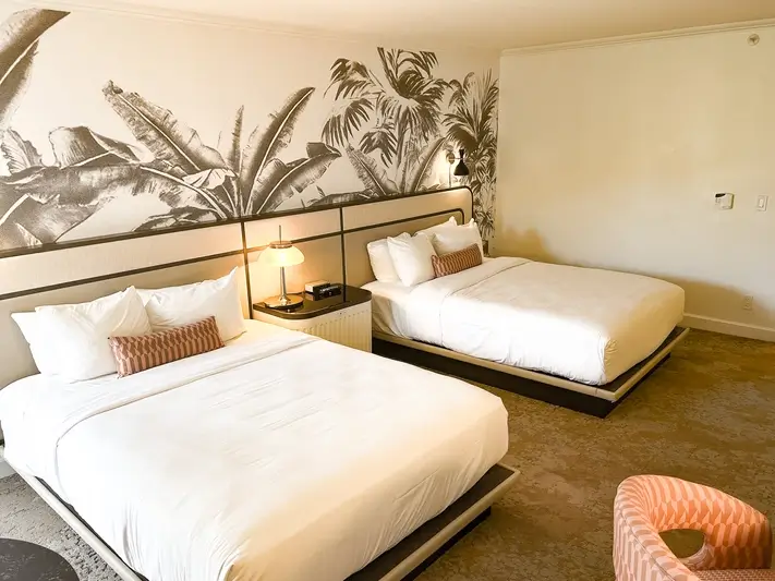 A well-lit hotel room featuring two queen-sized beds with crisp white linens and patterned accent pillows. Behind the beds, a large mural of black-and-white tropical palm leaves spans the wall. A small nightstand with a lamp sits between the beds, and the room is finished with neutral-toned carpeting.
