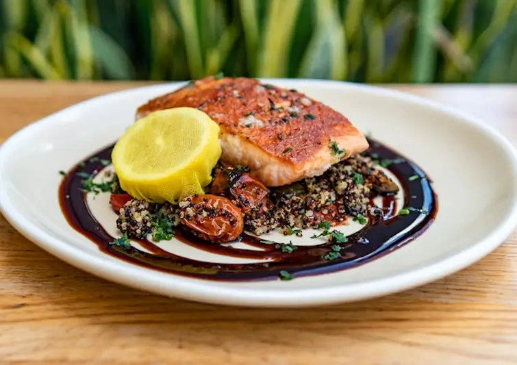 A gourmet plated meal featuring a seared salmon fillet resting on a bed of dark quinoa and roasted cherry tomatoes. The plate is decorated with a swirl of dark balsamic glaze and a garnish of fresh herbs. A bright yellow lemon half, wrapped in a thin yellow mesh, sits on the side.