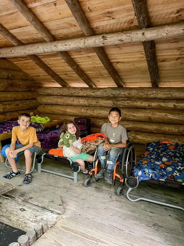 Three children are smiling inside a rustic log cabin. On the left, a boy in a yellow shirt sits on a small cot. In the middle, a girl in a white patterned shirt holds a green teddy bear while sitting on her bed. On the right, a boy in a grey t-shirt sits in his wheelchair. The room is filled with several small cots and colorful blankets under a slanted wooden ceiling.