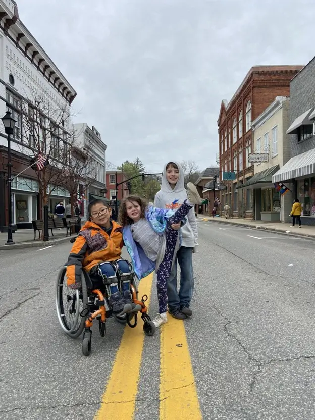 Three children pose playfully in the middle of a quiet town street marked with double yellow lines. A boy in a wheelchair wears an orange and blue jacket on the left. Beside him, a girl in a purple jacket strikes a pose with one leg kicked up high. A taller boy in a grey hoodie stands behind her. The street is lined with historic brick buildings under an overcast sky.