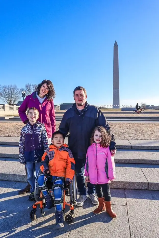 A full family portrait taken outdoors on a sunny day. Two adults and three children stand together on stone steps. The Washington Monument rises tall in the background against a crisp blue sky. Everyone is dressed in warm winter jackets, including a boy in a wheelchair in the front row and a girl in a bright pink coat.