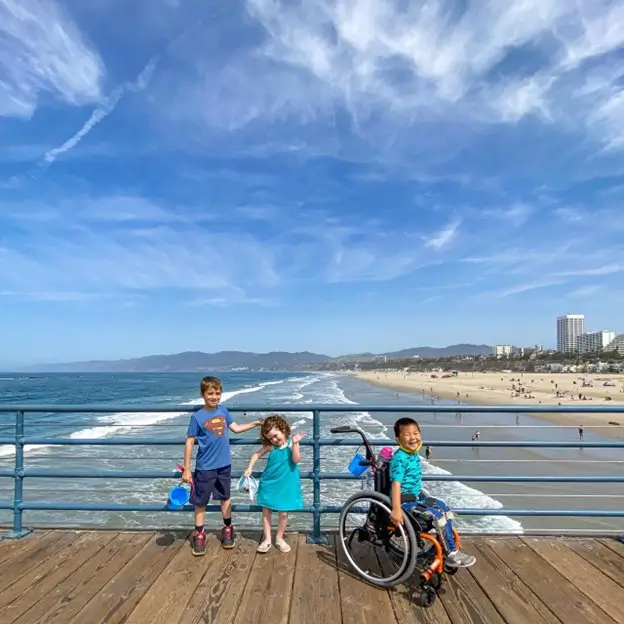 Three children stand together on a wooden pier under a vast blue sky with wispy clouds. To the left, a boy in a blue Superman shirt and dark shorts holds a blue bucket. In the center, a young girl in a teal dress waves with a smile. On the right, a boy sitting in a wheelchair wears a bright patterned blue shirt and grins at the camera. The ocean waves and a sandy coastline with distant buildings are visible in the background.