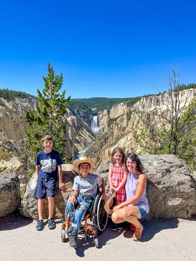 A family of four poses on a sunny day at a scenic overlook. A woman kneels on the right next to a young girl in a pink plaid dress. In the center, a boy in a straw hat sits in a wheelchair, and to the left, another boy stands near a pine tree. They are positioned in front of a massive canyon with a large waterfall cascading in the distance under a clear blue sky.