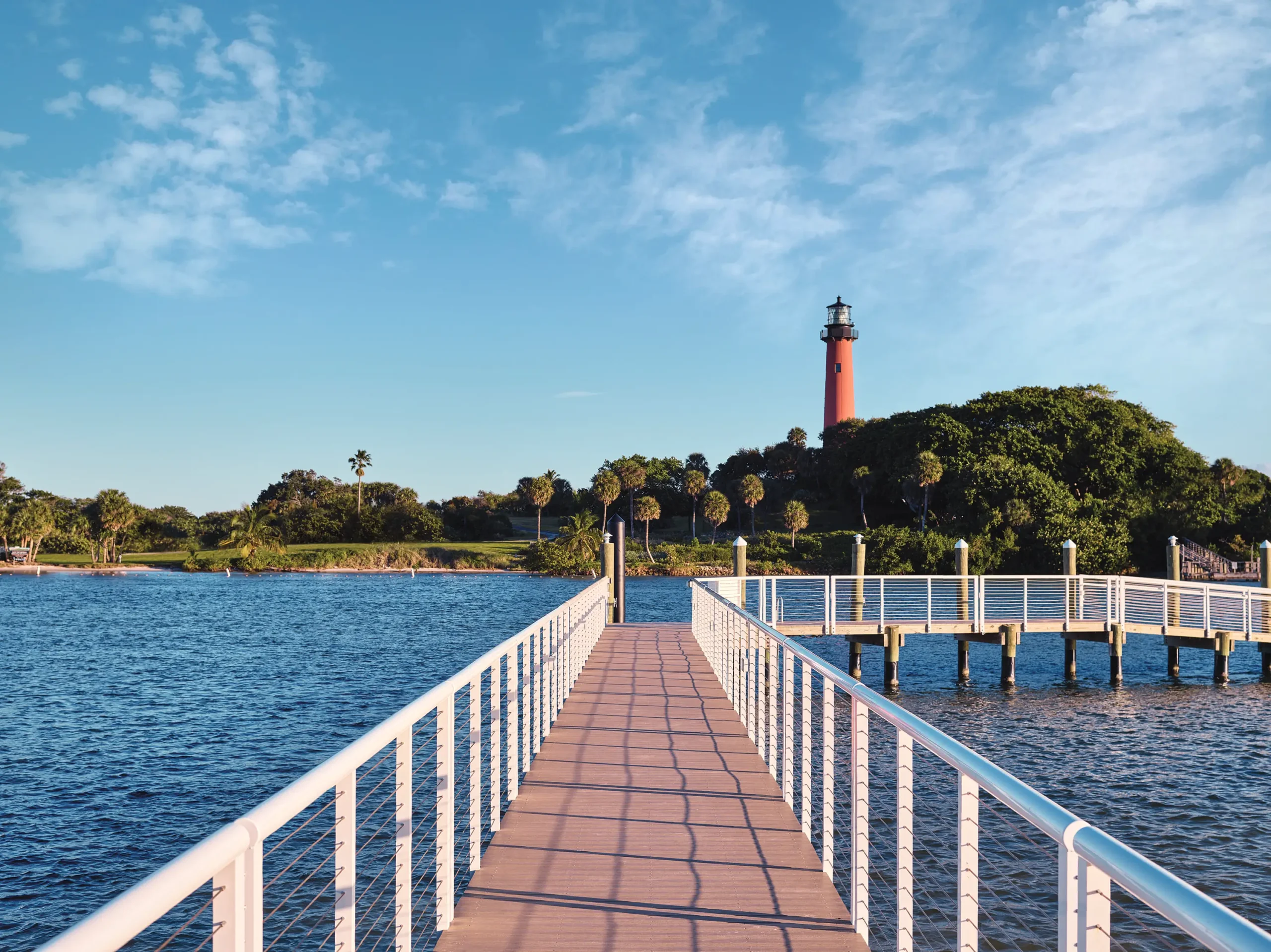 A wide-angle shot from a wooden boardwalk with white metal railings extending over deep blue water toward a lush, green shoreline. In the distance, a tall, bright red lighthouse stands prominently among a cluster of palm trees and dense foliage. The sky above is a clear light blue with soft, scattered white clouds, and the scene is illuminated by the warm, golden light of late afternoon.