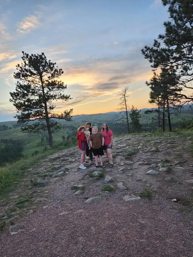 A wide shot of five people standing on a rocky, dirt hilltop at sunset. The sky is a blend of soft blue and pale yellow light behind rolling green hills. Two tall, thin pine trees frame the group, who are dressed in casual summer clothes.