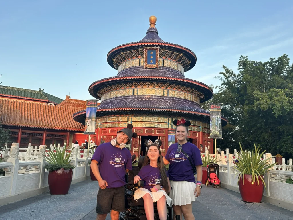 Three people pose in front of a traditional Chinese pagoda (The Hall of Prayer for Good Harvests). They are all wearing matching purple t-shirts that say "Cure SMA." The woman in the center is in a power wheelchair wearing sequined mouse ears.
