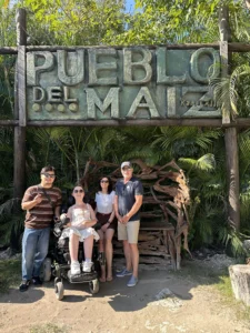 A group of four people poses under a large wooden sign that reads "Pueblo Del Maíz." The woman in the power wheelchair is at the front left, surrounded by three companions amidst lush tropical greenery.