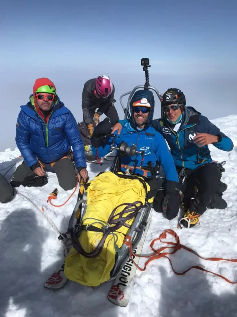 Three climbers pose for a celebratory photo on a snowy summit. The climber in the center sits in an adaptive sled, flanked by two teammates in high-altitude gear. They are all smiling, with a thick layer of clouds visible in the valley below them.