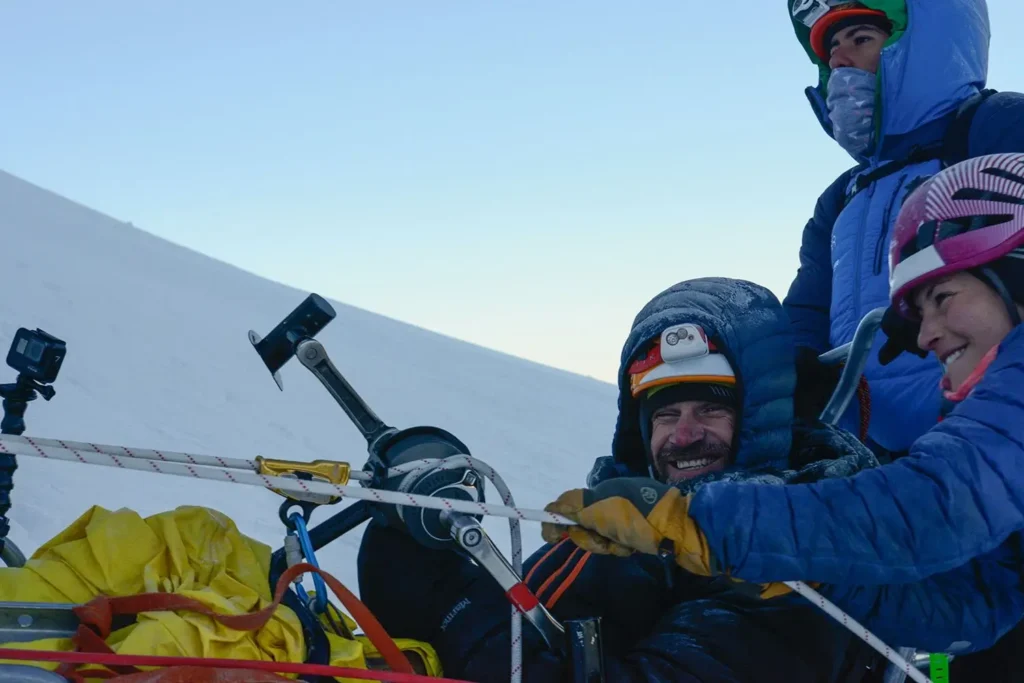 A close-up of three climbers on a snowy mountain. In the center, a man in a blue hooded jacket and headlamp smiles broadly despite the harsh conditions. A person in a blue jacket and pink helmet is to his right, and another climber in a blue hood is behind them. They are surrounded by climbing ropes and rigging.