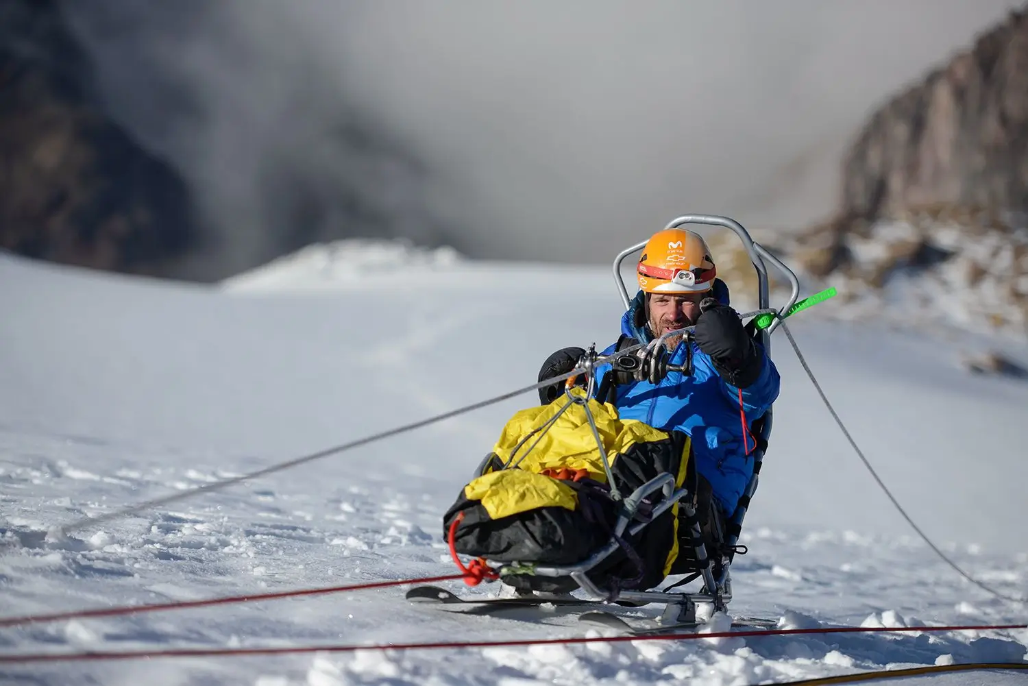 A mountaineer wearing an orange helmet and blue jacket sits in a specialized sit-ski or adaptive sled on a snow-covered mountain. He is holding onto ropes, and the sled is rigged with several safety lines extending across the bright, white landscape.