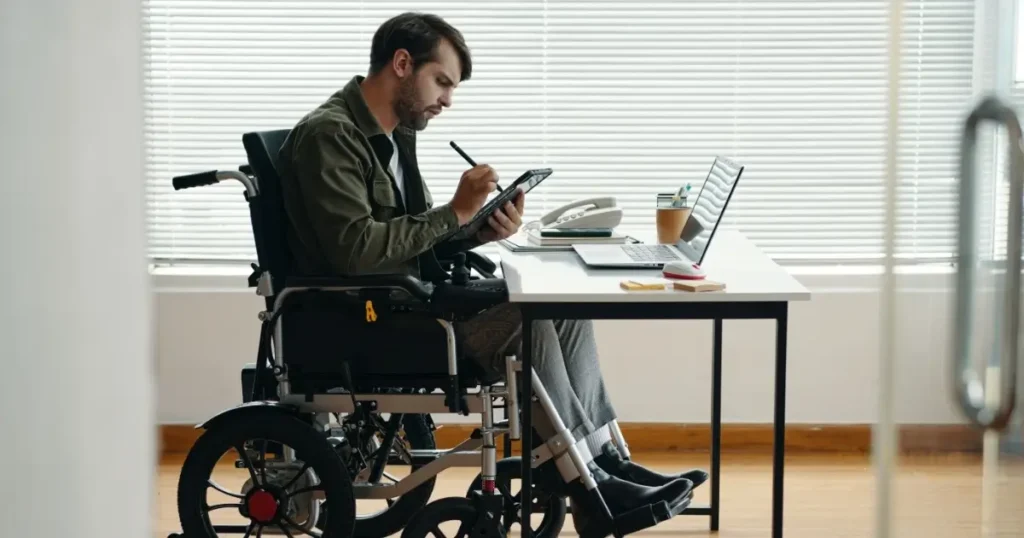 A man using a wheelchair works at a white desk in a bright office, using a stylus on a digital tablet next to a laptop and telephone.