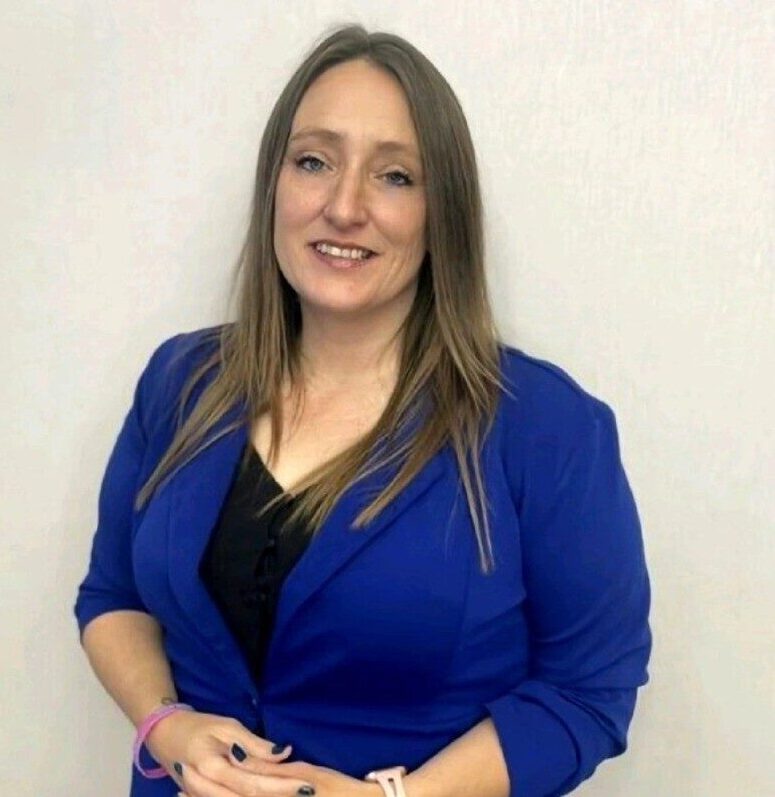 A professional portrait of a woman with long light-brown hair, smiling at the camera. She is wearing a bright blue blazer over a black top and standing against a plain white background.