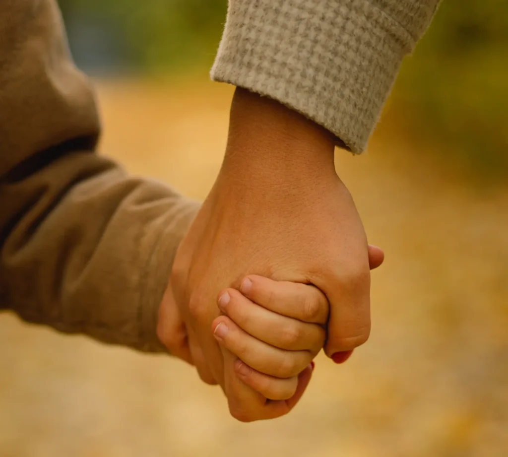 A close-up, vertical shot of an adult's hand firmly and gently holding a young child's hand. The hands are centered in the frame, with the adult wearing a light-colored, textured sleeve and the child in a tan or brown ribbed sleeve. The background is a soft, out-of-focus golden yellow, suggesting a path covered in autumn leaves.