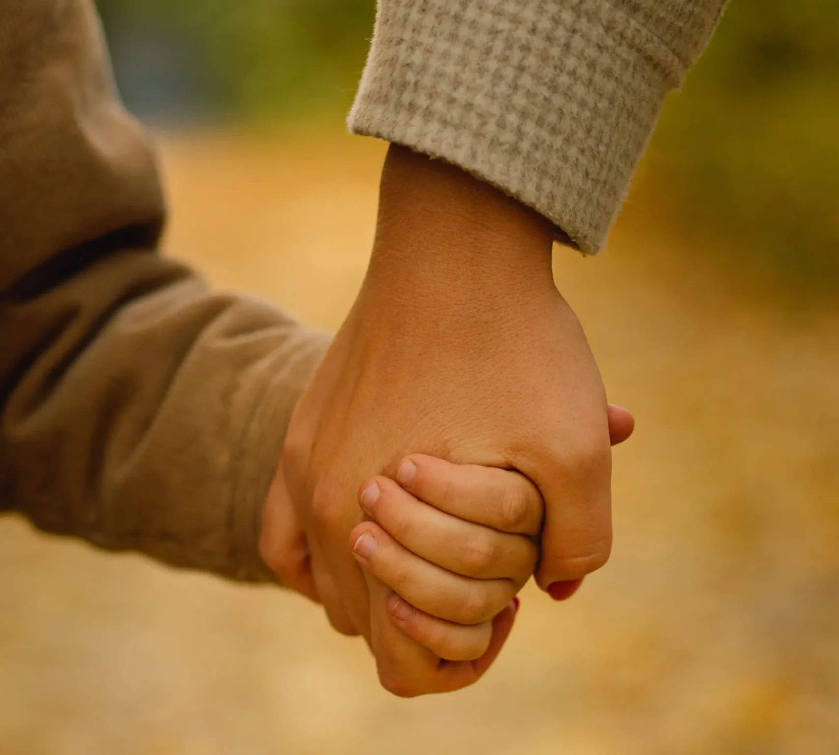 A close-up, vertical shot of an adult's hand firmly and gently holding a young child's hand. The hands are centered in the frame, with the adult wearing a light-colored, textured sleeve and the child in a tan or brown ribbed sleeve. The background is a soft, out-of-focus golden yellow, suggesting a path covered in autumn leaves.