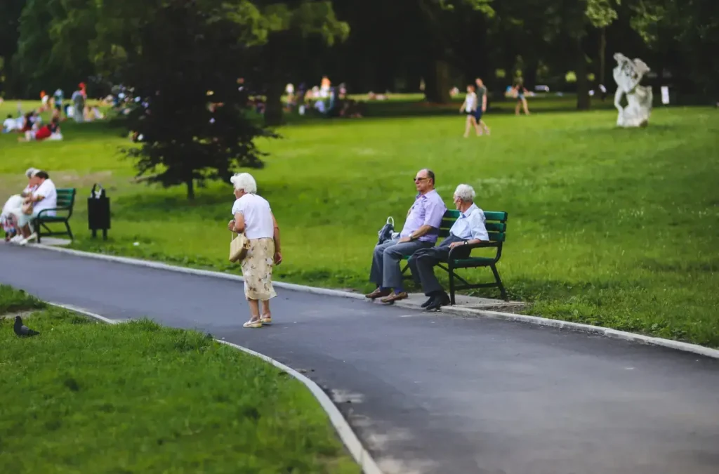 Elderly people relaxing on green park benches along a paved walking path on a sunny afternoon.