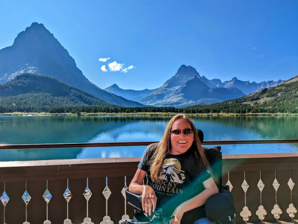A woman in a wheelchair smiling on a wooden balcony at Glacier National Park, featuring a crystal-clear blue lake and dramatic mountain peaks under a bright sky.