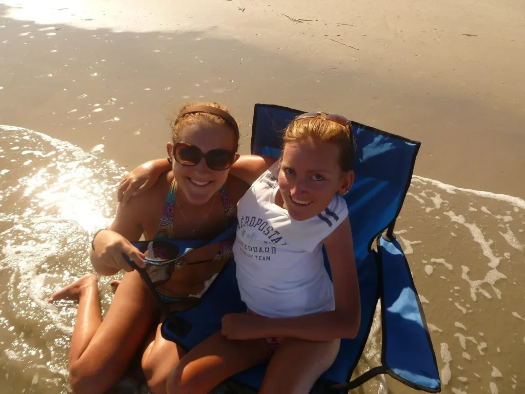 Two women smiling and embracing while sitting in a blue beach chair at the edge of the ocean tide on a sunny beach in Hilton Head.